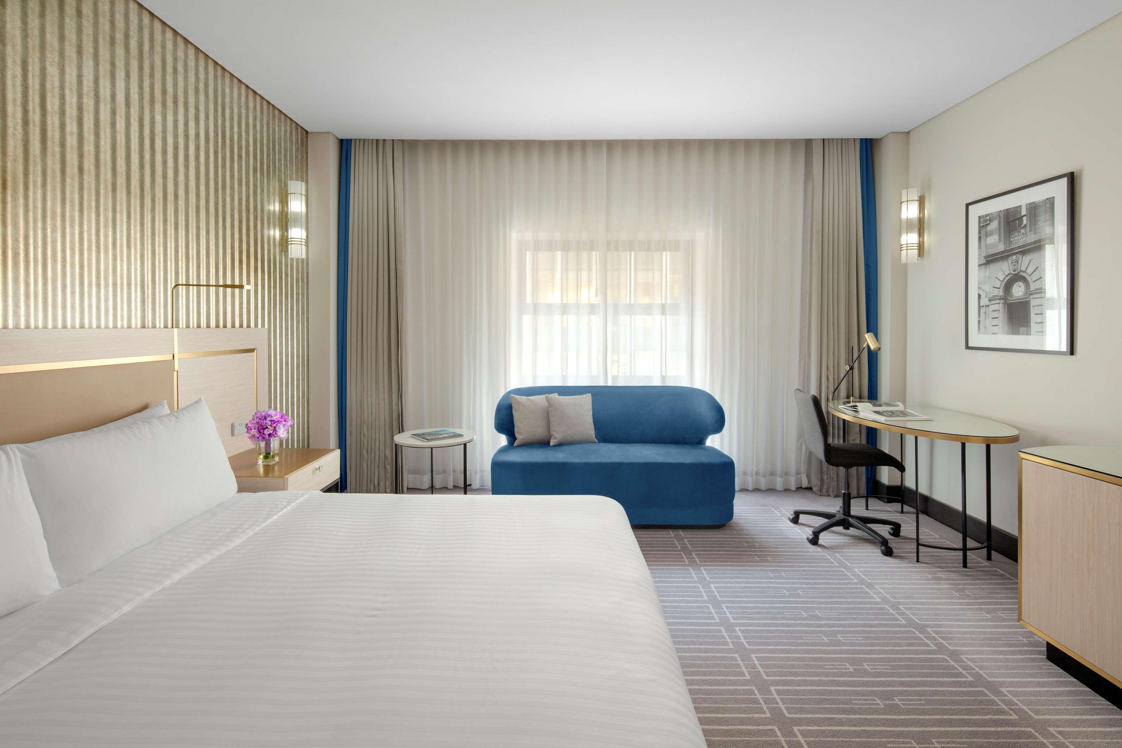 Radisson Blu Plaza Hotel Sydney Zewnętrze zdjęcie The photo shows a modern hotel room featuring a large bed with white linens. There's a small table with a flower vase next to the bed. A blue sofa is positioned against one wall, and a desk with a chair is situated nearby. The room has soft, diffused