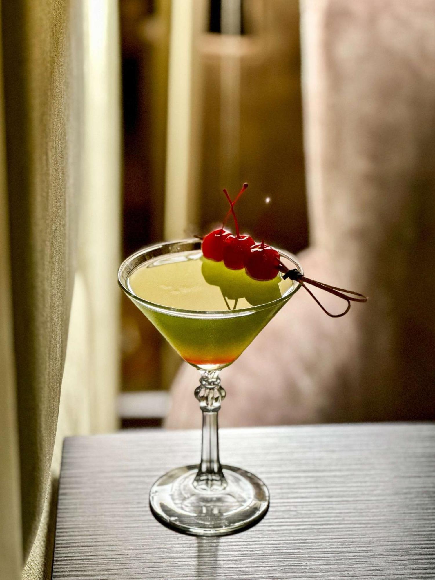 Radisson Blu Plaza Hotel Sydney Zewnętrze zdjęcie The image shows a cocktail in a classic martini glass. The drink has a vibrant green color and is garnished with two yellow-green fruits (possibly a melon or similar) and three cherries on a skewer. The background appears blurred, creating a soft foc