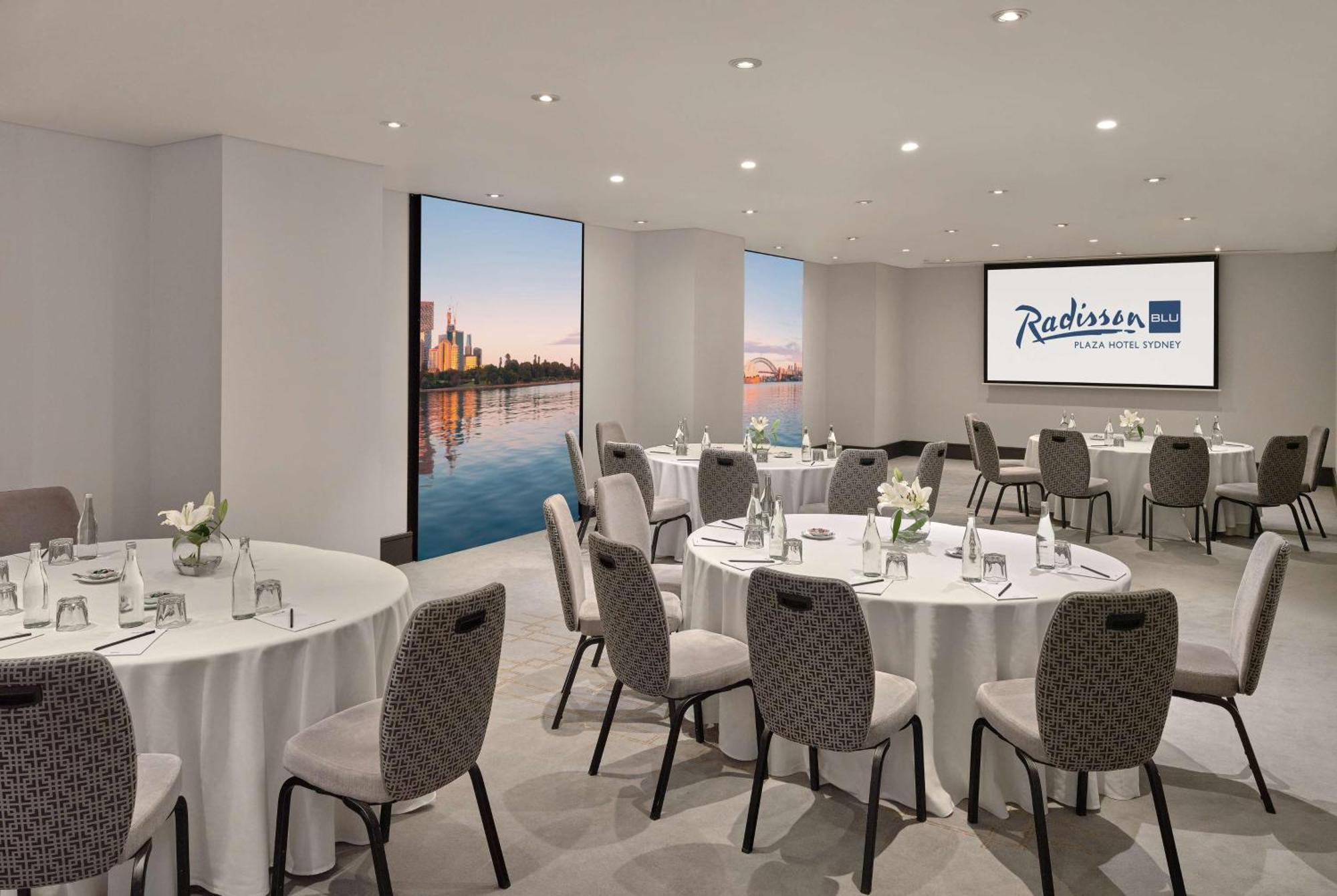 Radisson Blu Plaza Hotel Sydney Zewnętrze zdjęcie The photo shows a modern meeting room at the Radisson Hotel in a city. The room features several round tables set with white tablecloths and gray upholstered chairs. There are small flower arrangements on each table, creating a welcoming atmosphere. 