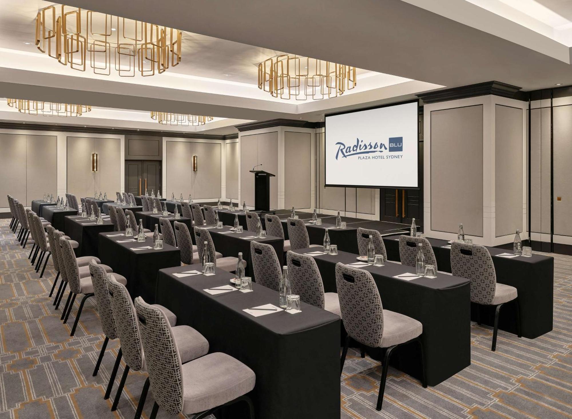 Radisson Blu Plaza Hotel Sydney Zewnętrze zdjęcie The photo shows a conference or meeting room set up in a hotel, likely a Radisson. The room features several rows of tables covered in black tablecloths, each with a chair beside it. Water glasses are placed on each table, and there appears to be a l